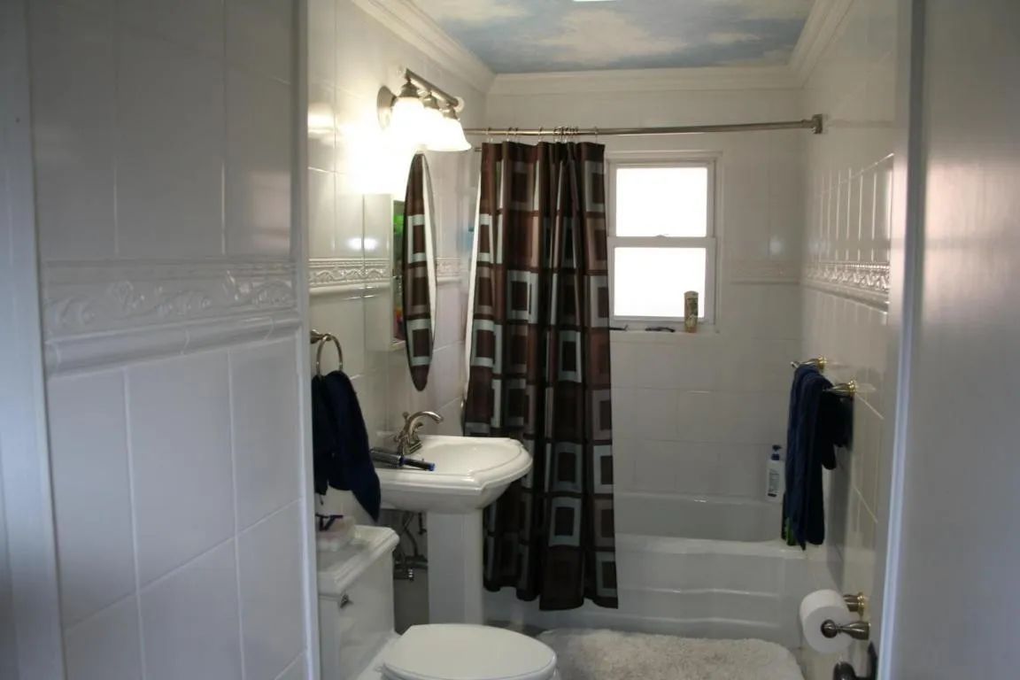 A bathroom with white tile walls, a pedestal sink, a toilet, a shower with a brown geometric curtain, and a cloud ceiling.