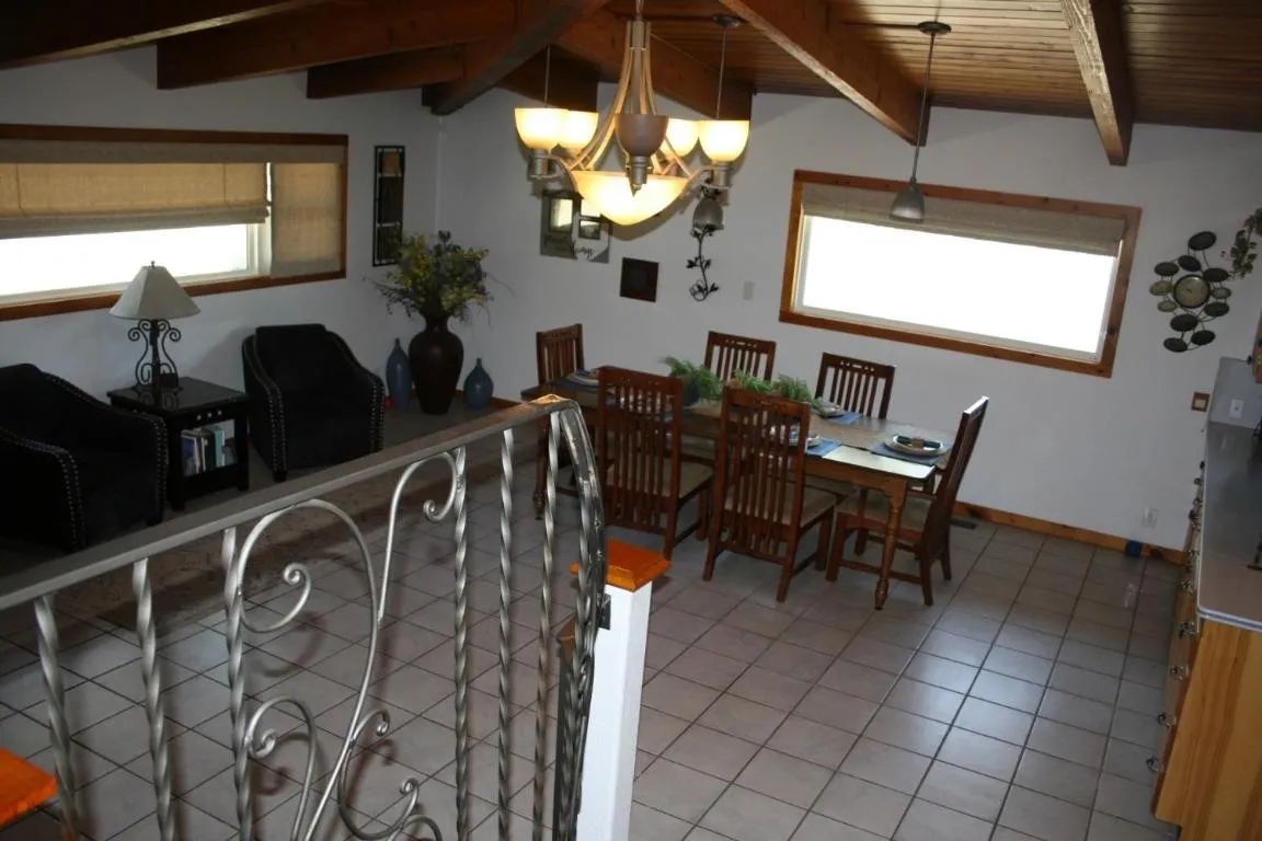 A dining area with a wooden table, chairs, and a chandelier, viewed from behind a decorative metal stair railing.