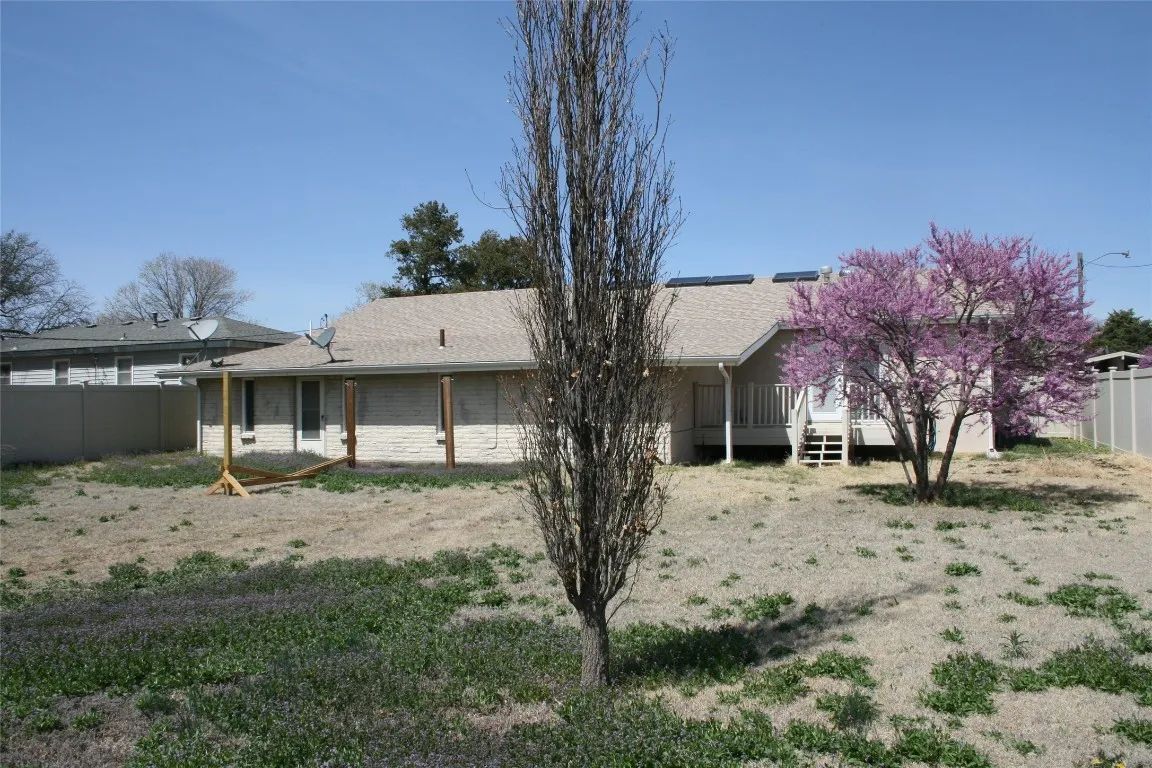 A backyard view of a light-colored house with a tall, thin tree in the foreground and a blooming purple tree to the right.