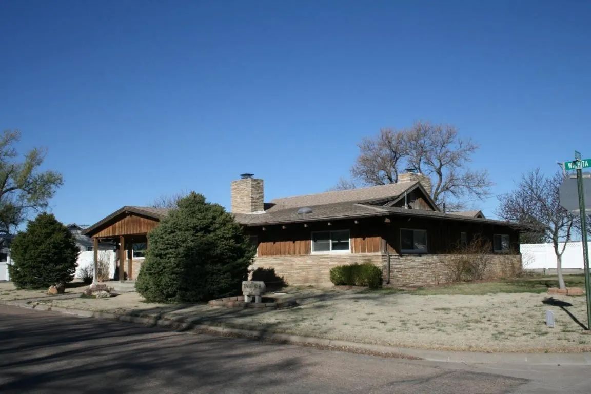 A tan brick and wood-sided ranch-style house with a chimney and large evergreen bushes on a clear day.