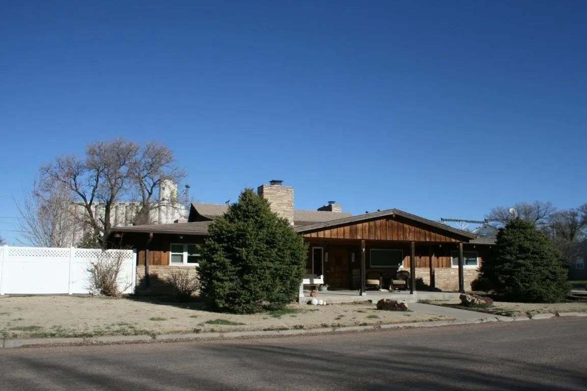 A low-slung, wood-sided house with a porch and brick chimney sits under a clear blue sky, next to a white privacy fence.