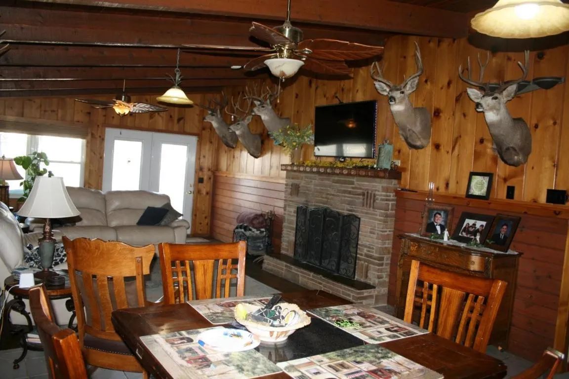 Rustic living room with wood paneled walls, stone fireplace, mounted deer heads, and a dining table in the foreground.