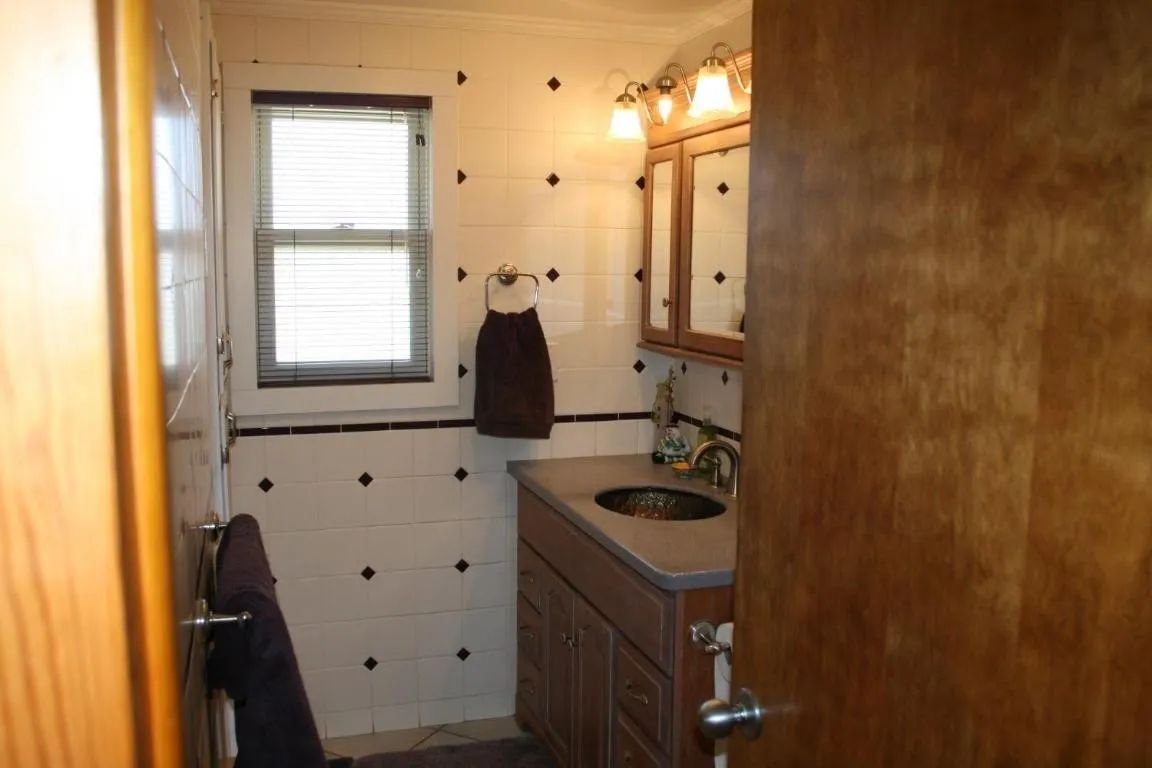 A small bathroom featuring white tiled walls with dark accents, a wood vanity, a medicine cabinet, and a window.
