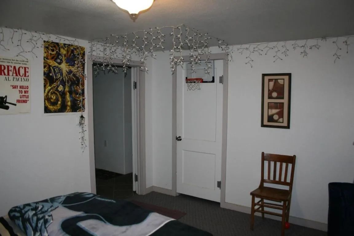 A room with a bed, a chair, and wall decorations, including a poster and lights, with an open doorway in the center.