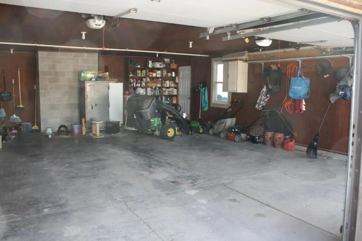 A cluttered two-car garage with concrete floors, a riding mower, shelving units, and various items hanging on the walls.