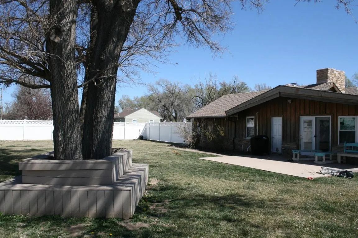 A large tree with a tiered stone planter box stands in a residential yard beside a brown house with a patio.