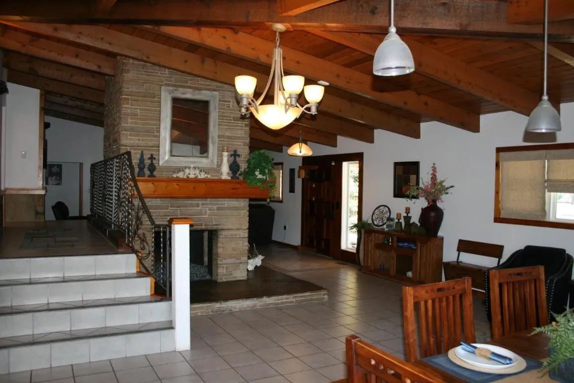A vaulted, wood-beamed living room with a stone fireplace, tile floors, a dining area, and warm, overhead lighting.