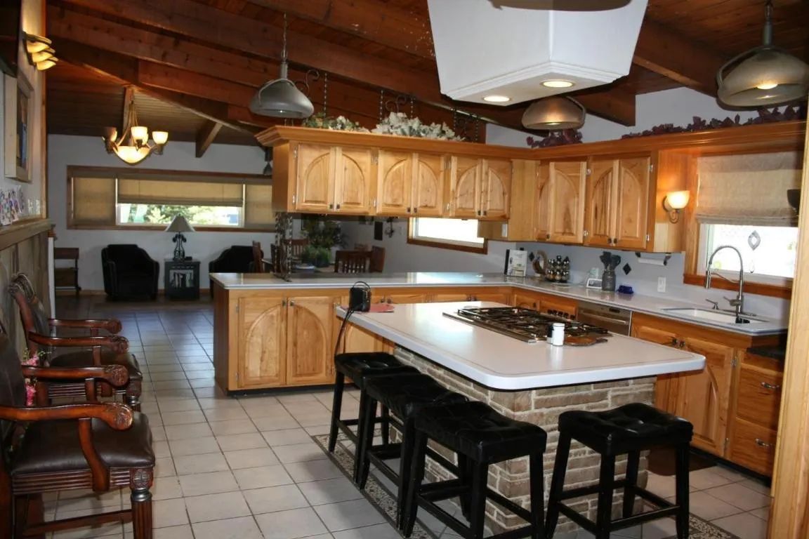 A kitchen with light wood cabinets, a brick-based island with black stools, and a view into a living area with a fireplace.