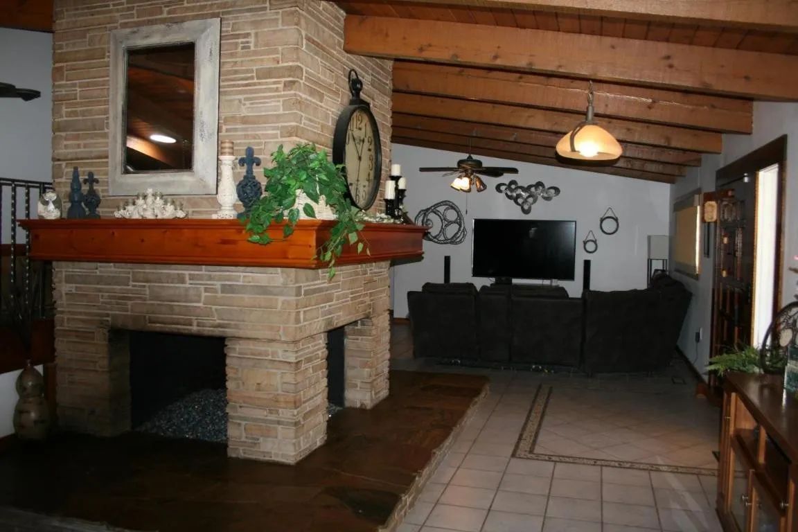 A living room with a large, central stone fireplace, wooden beams, a flat-screen TV, and dark seating.