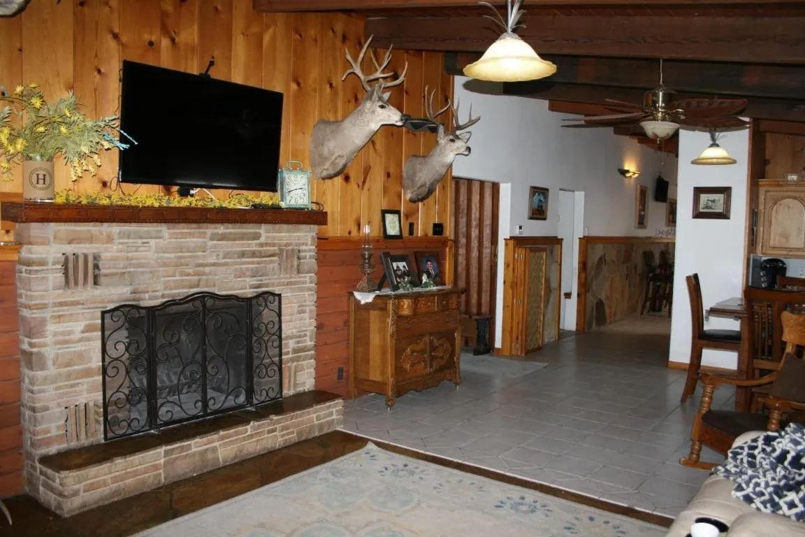 A rustic living room featuring a stone fireplace, wood-paneled walls, two deer mounts, and an open dining area.