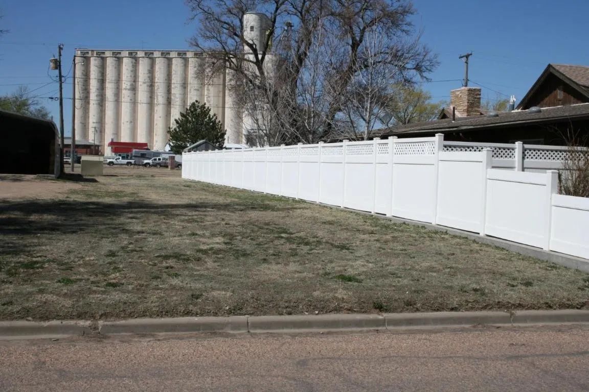 A white vinyl fence borders a dry grass lot in front of tall industrial grain silos under a clear blue sky.