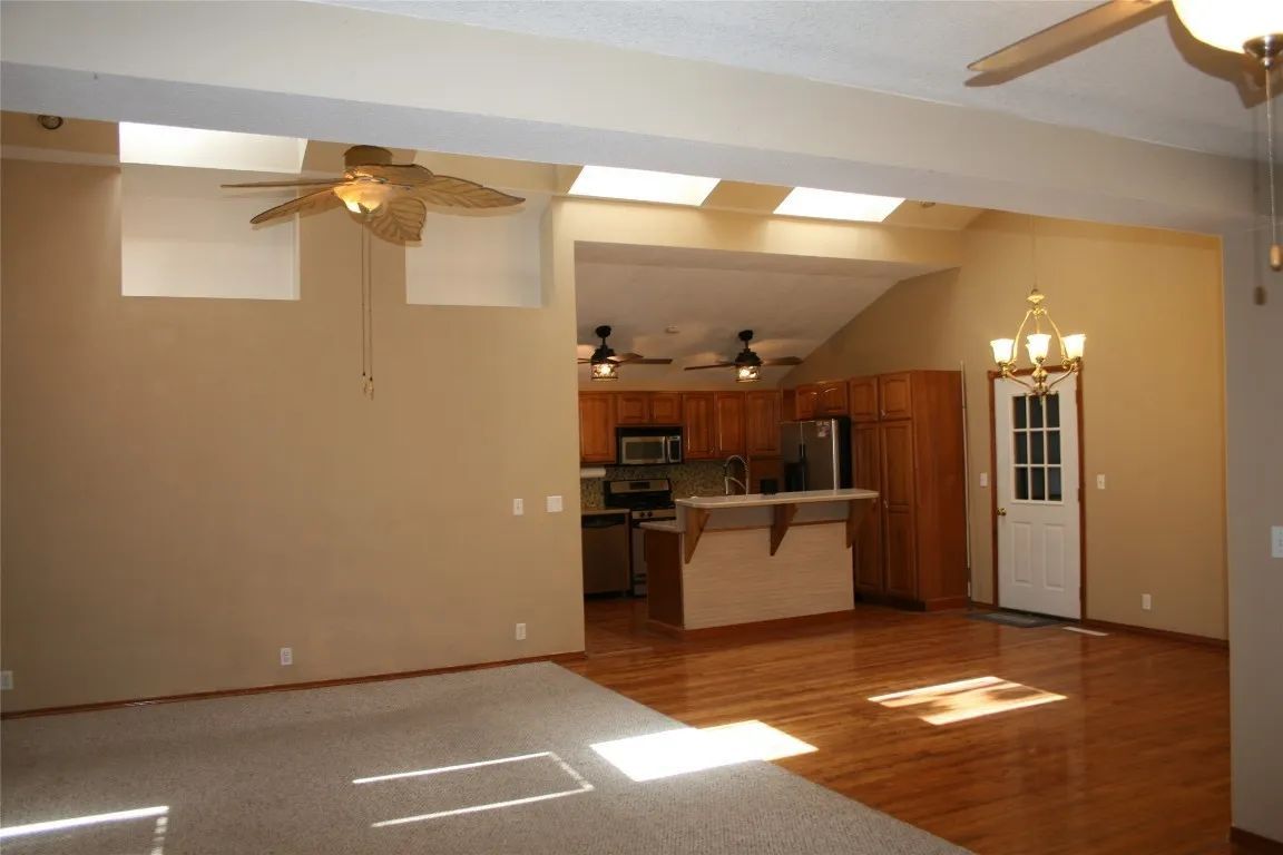 An open-concept living area with a carpeted foreground, wooden floors in the kitchen, and bright skylights above.