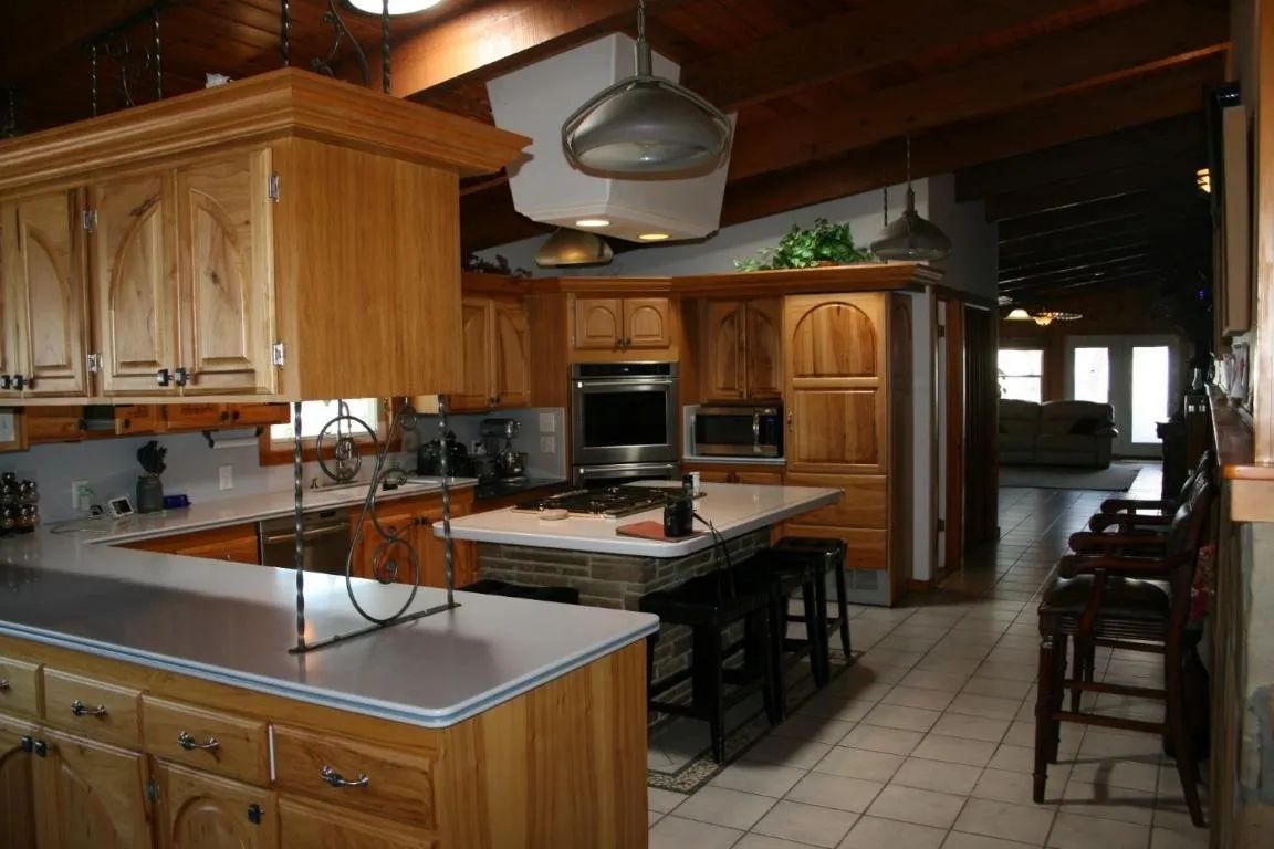A kitchen with natural wood cabinets, a stone-base island with black stools, and a tile floor, viewed from an entryway.