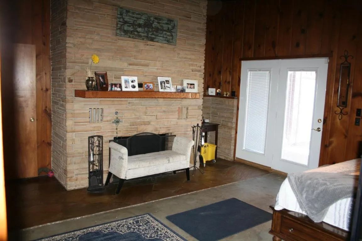 A stone fireplace with a wooden mantel, a white upholstered bench, and double glass doors in a wood-paneled room.