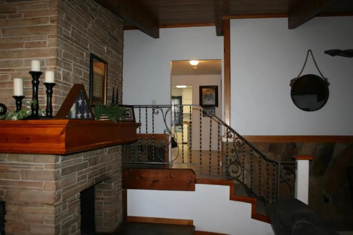 A stone fireplace with a wooden mantle and candles, next to a staircase with ornate iron railings leading to a doorway.
