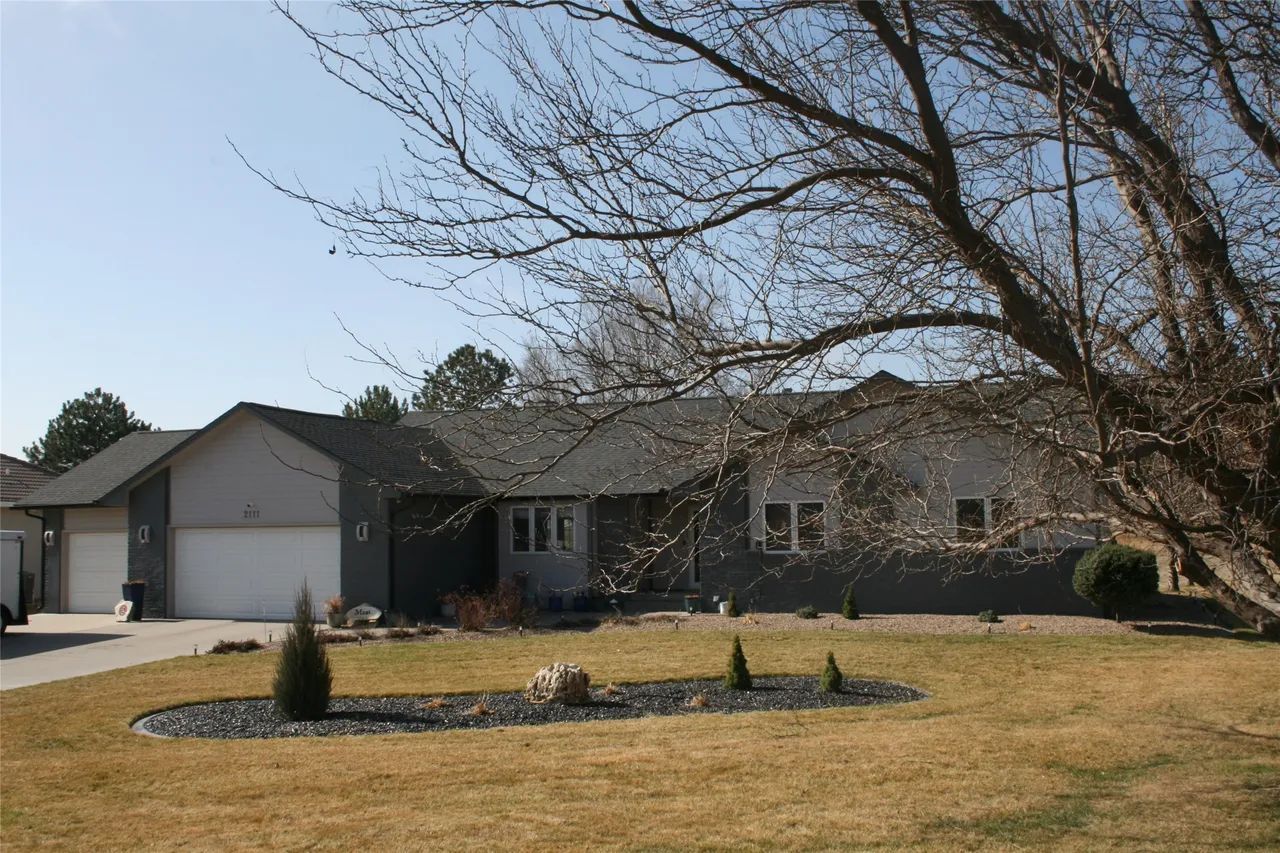 A single-story residential home with a two-car garage, grey siding, and a large tree in the front yard.