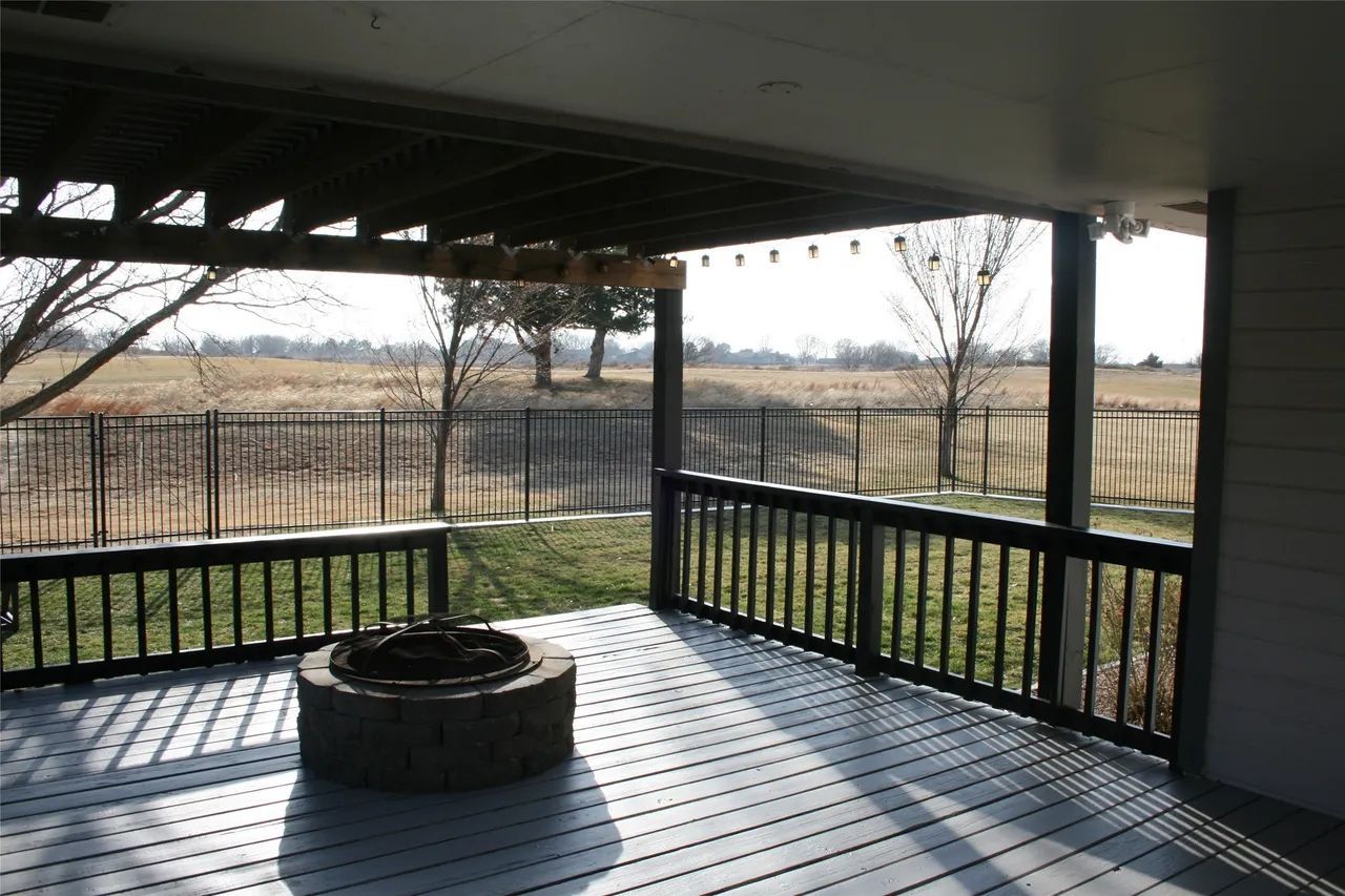 A gray wooden deck with a stone fire pit, black railings, and a view of a grassy field beyond a metal fence.
