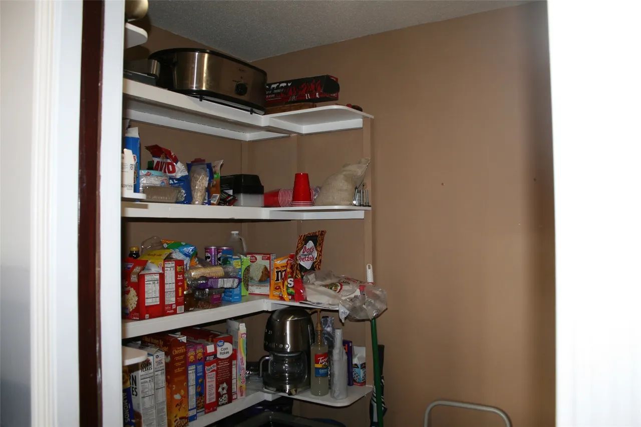 A pantry closet with shelves holding various food items, snacks, a slow cooker, a coffee maker, and cleaning supplies.