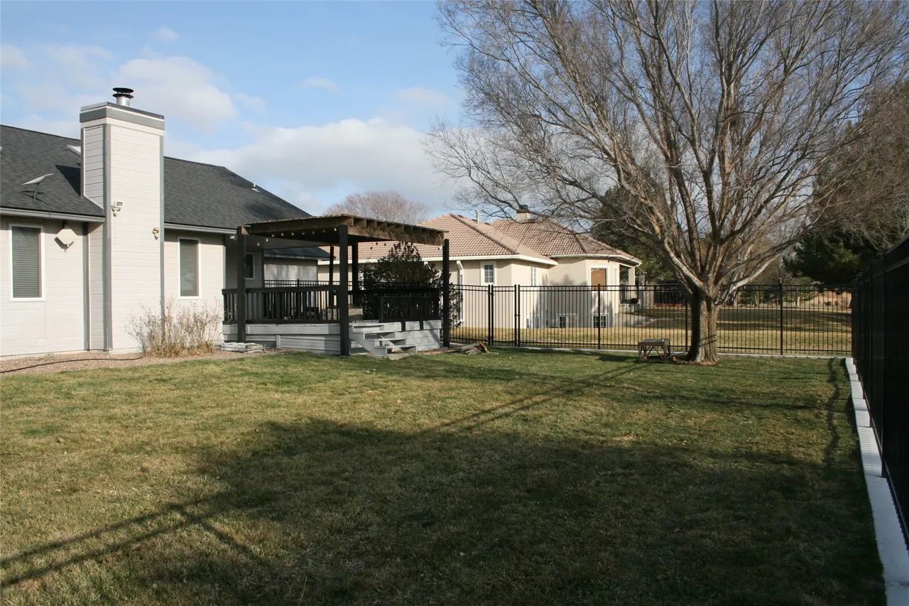 A suburban backyard with a grassy lawn, a patio with a dark pergola, a white brick house, and a fence on a sunny day.