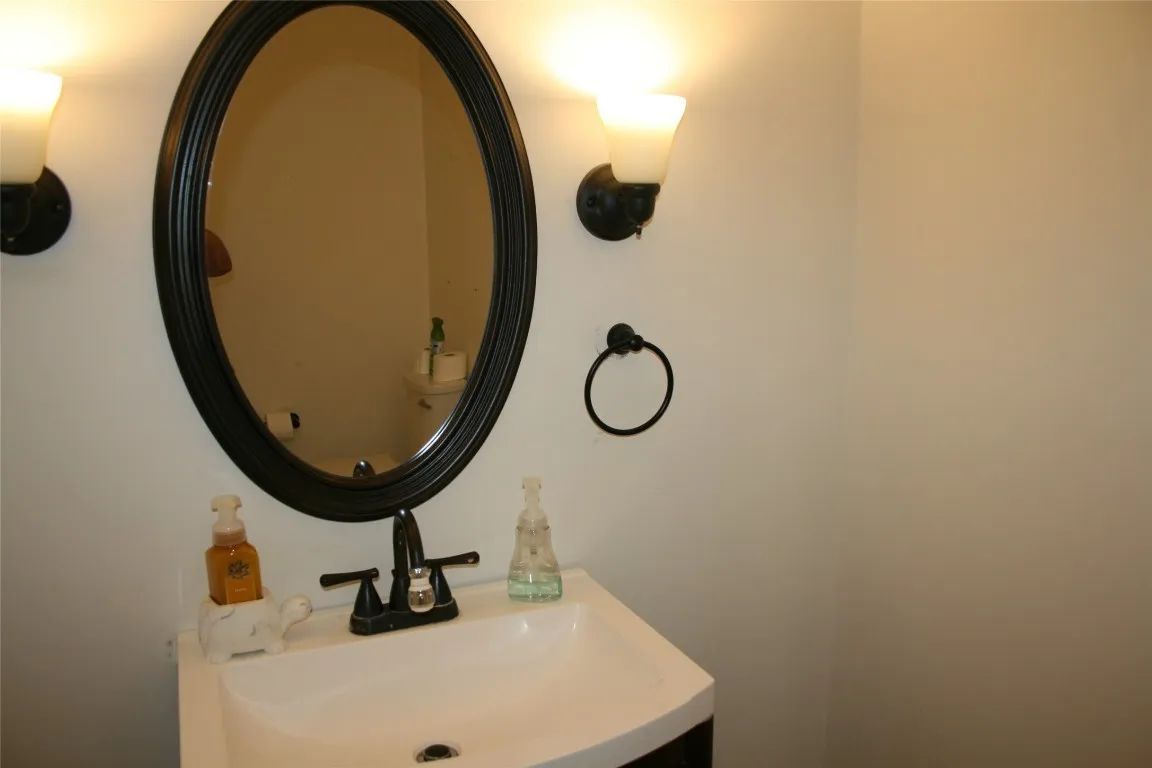 A white bathroom sink with a black faucet, oval mirror, two lights, and hand soap, all mounted on a plain white wall.