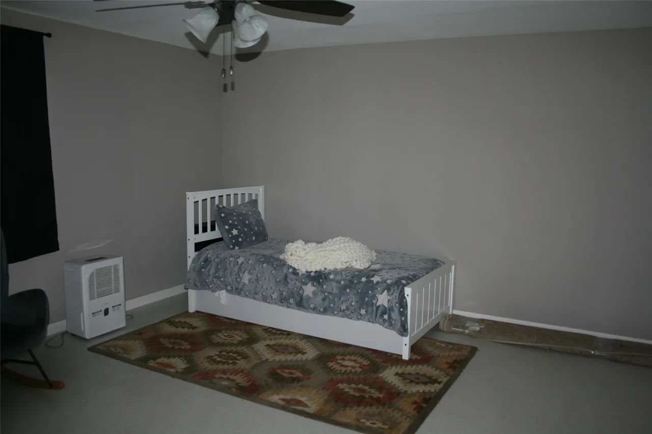 A white single bed with a patterned gray blanket sits on a brown area rug in a room with gray walls and a ceiling fan.