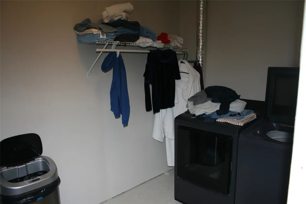 A laundry room with a trash can, a shelf holding folded clothes, hung garments, and dark washer and dryer units.