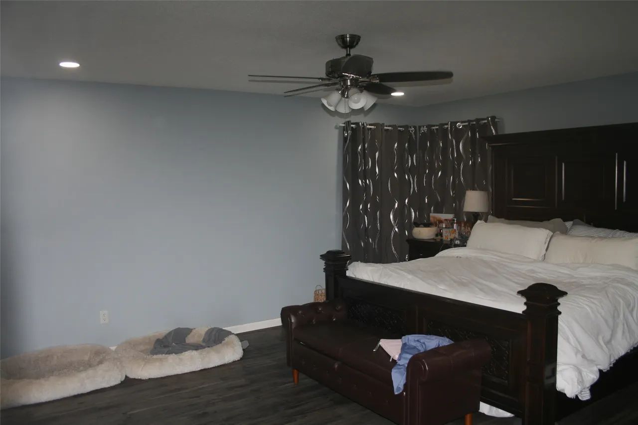 A bedroom with light gray walls, a dark wood bed with white bedding, a brown leather bench, and dog beds on the floor.