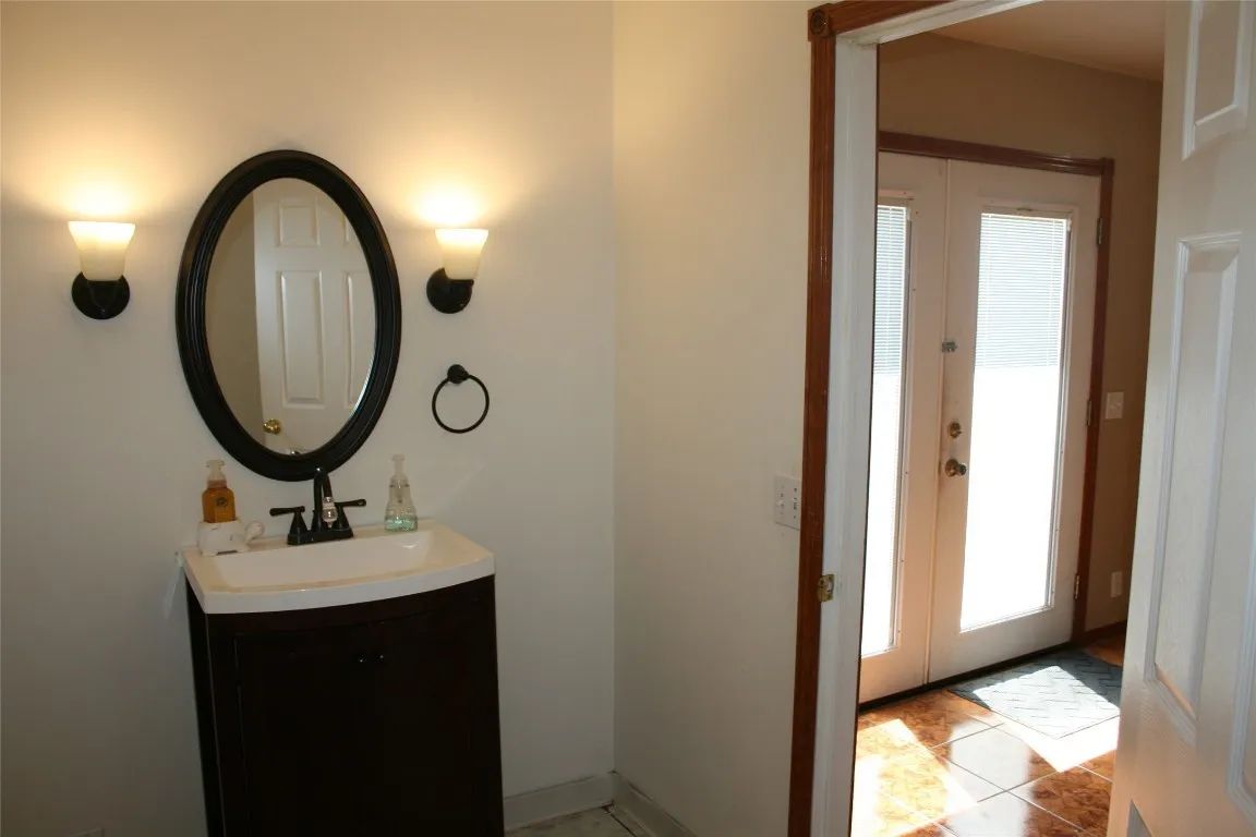 A bathroom vanity with an oval mirror and two wall lights, next to an open doorway leading to a brightly lit entryway.