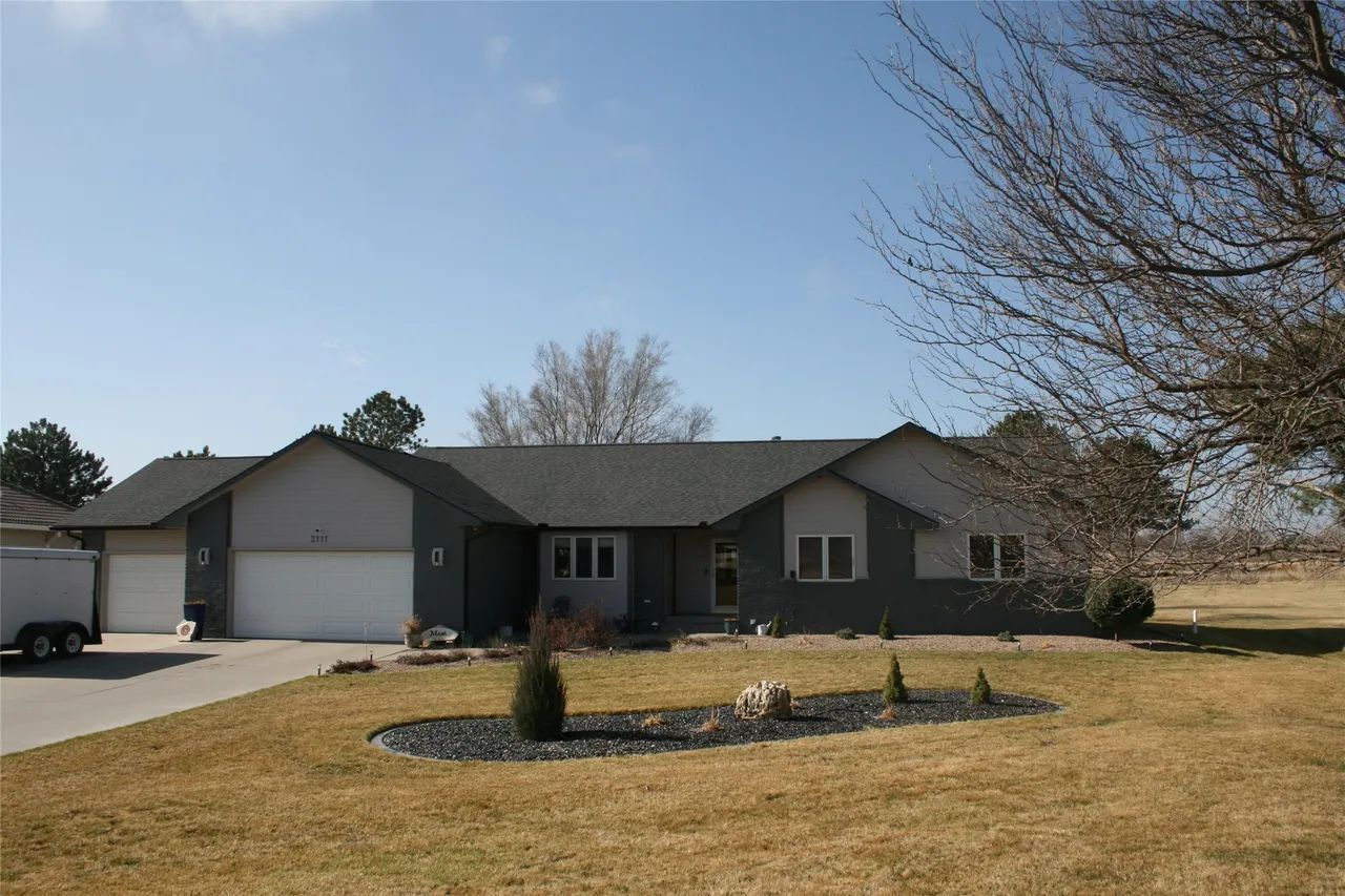 A wide shot of a gray, single-story house with a three-car garage, a paved driveway, and a dormant lawn on a sunny day.