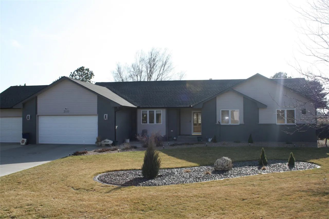 A single-story suburban house with a gray exterior, white garage doors, and a landscaped front yard with a rock bed.