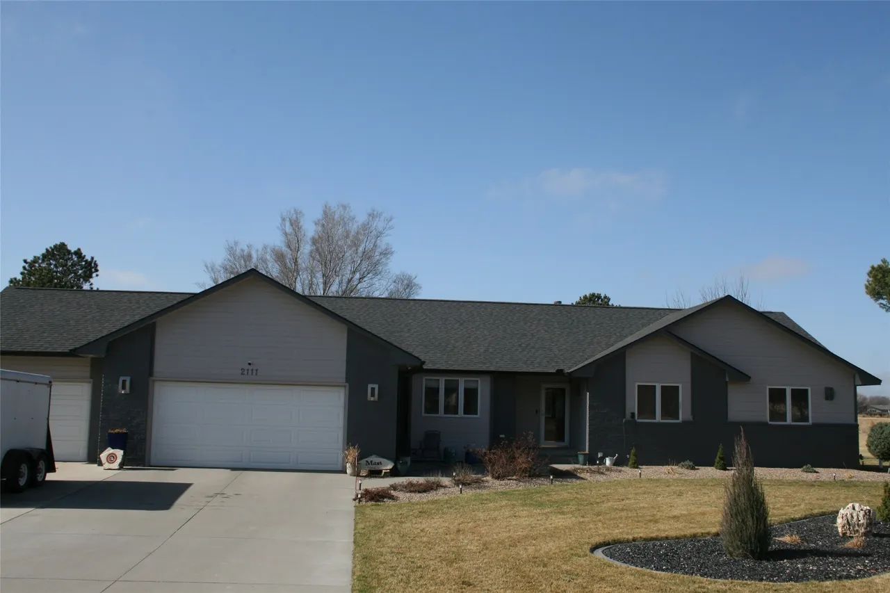 Single-story, gray ranch-style house with white garage doors and a wide driveway under a clear blue sky.