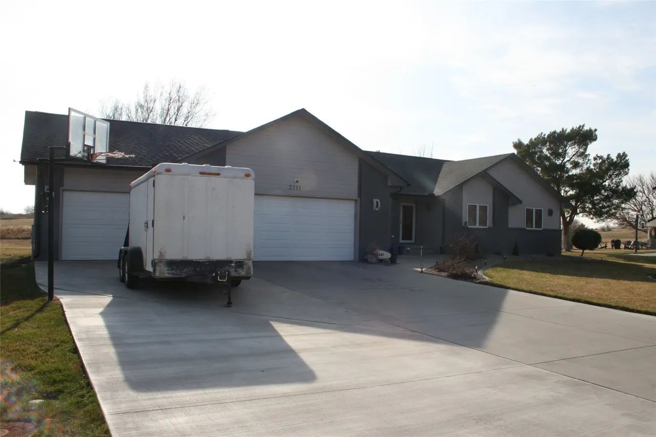 A suburban house with a multi-car garage, a parked cargo trailer in the driveway, and a basketball hoop.