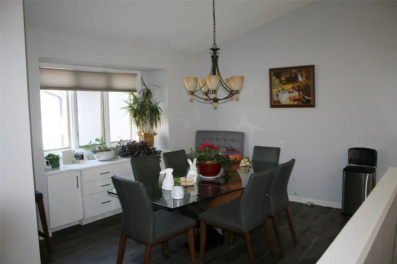 A dining room featuring a glass table with four grey chairs, a white side cabinet, a plant, and a hanging chandelier.