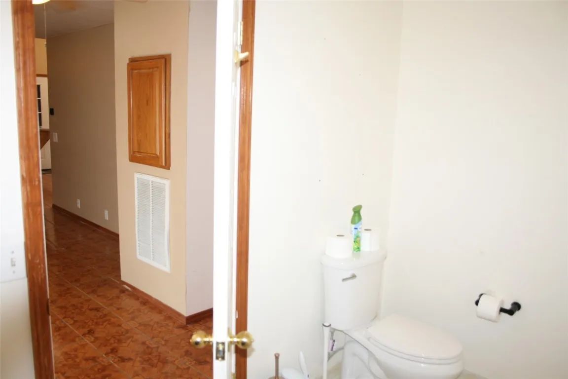 A bathroom with a toilet and toilet paper holder, viewed from a hallway with brown tile floors and beige walls.