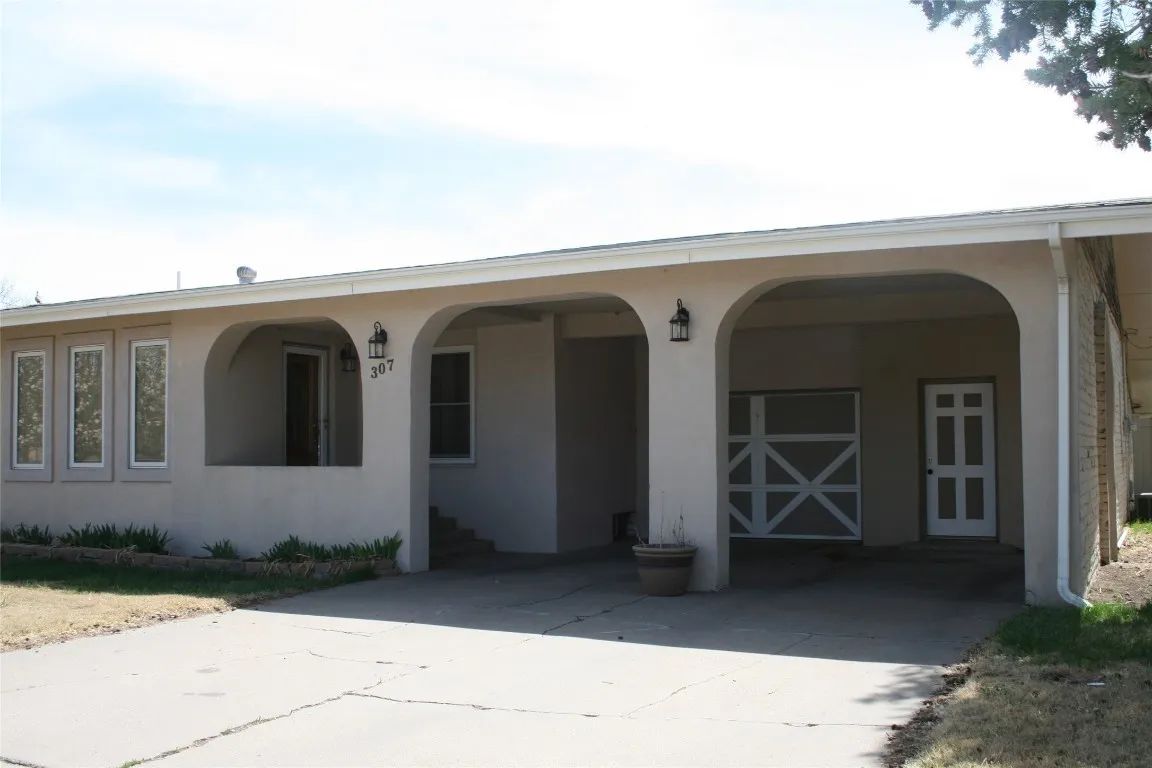 A beige, single-story house featuring a large, arched carport and a paved driveway under a bright sky.