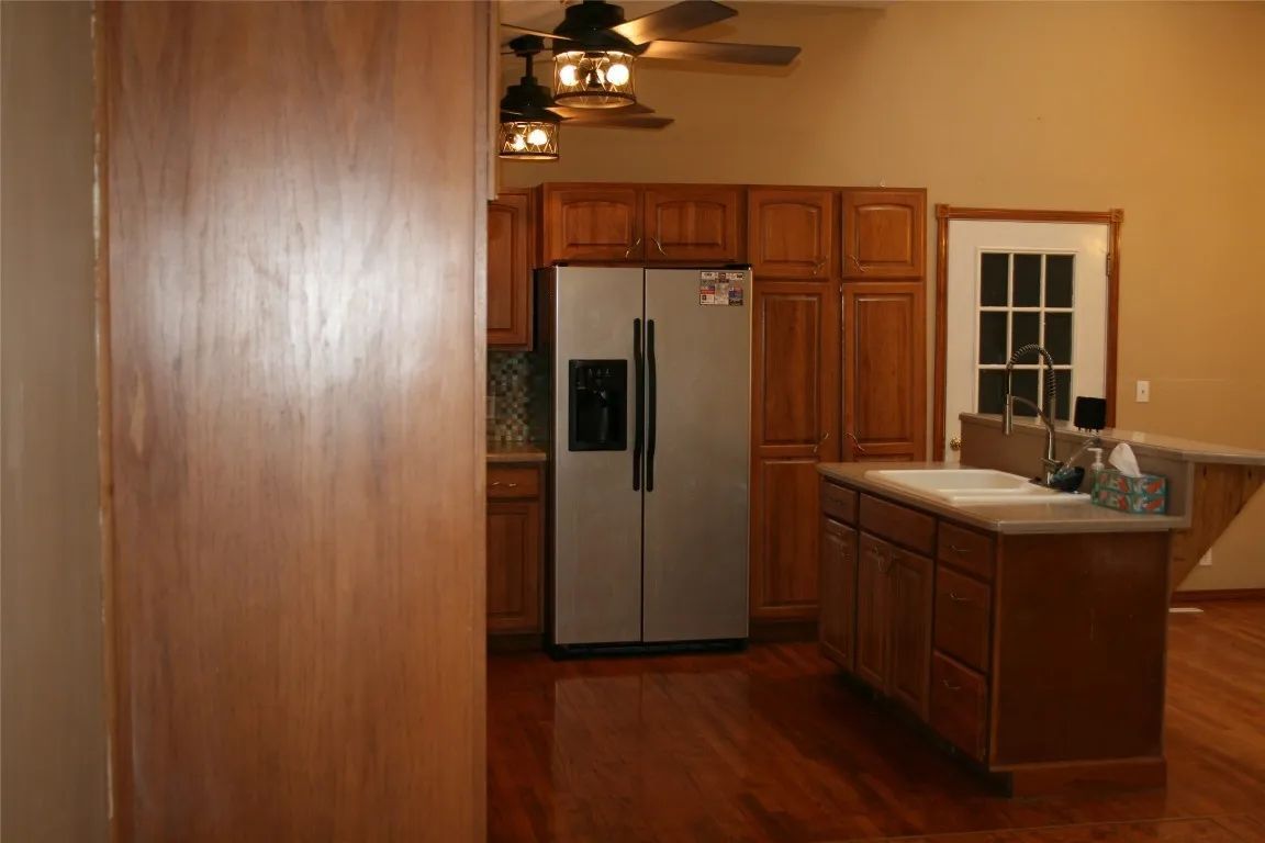 A kitchen with wood cabinets, a stainless steel refrigerator, a center island with a sink, and dark hardwood floors.