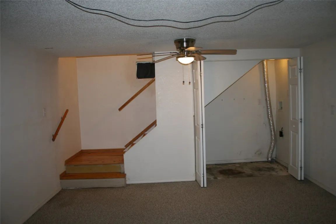 A carpeted basement room with wooden stairs leading up and a small, open storage alcove containing a metal vent.