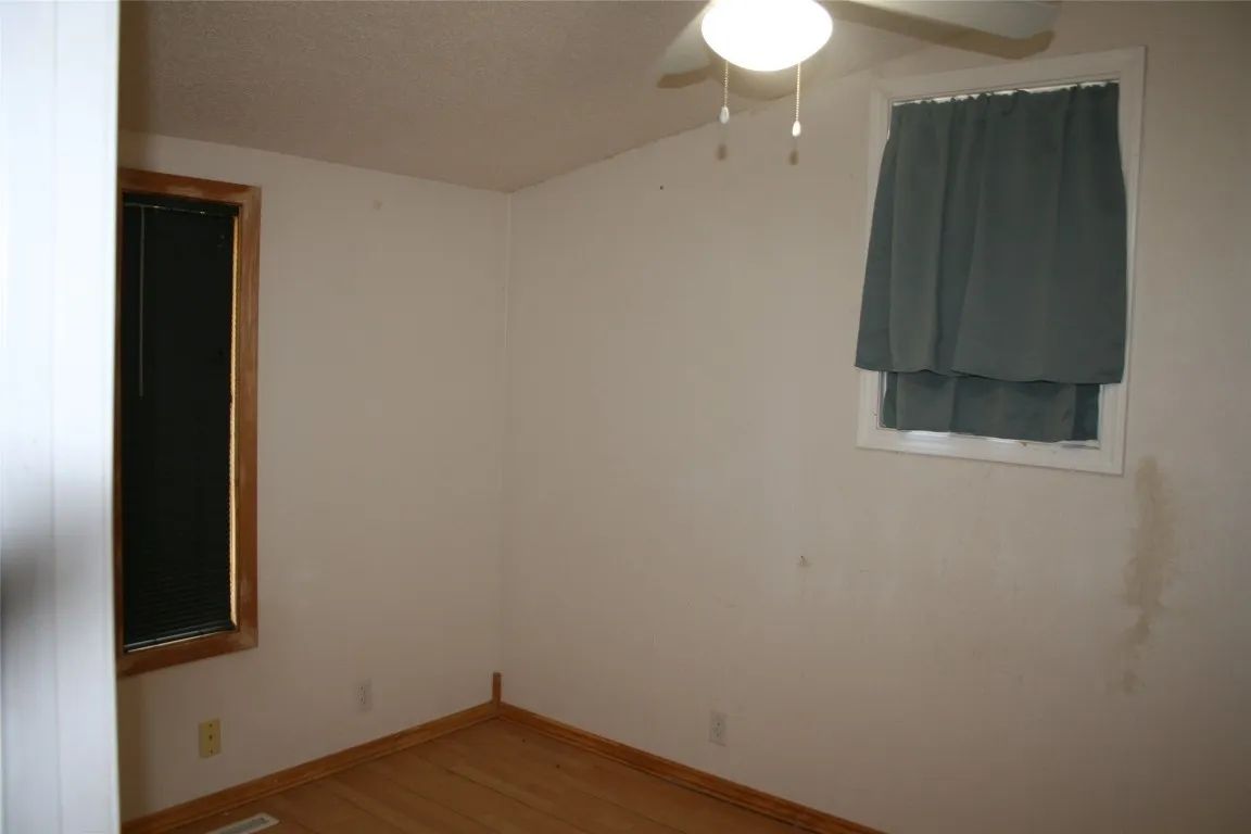 An empty room with beige walls, wood-tone trim, a window with grey curtains, and a ceiling fan light fixture.