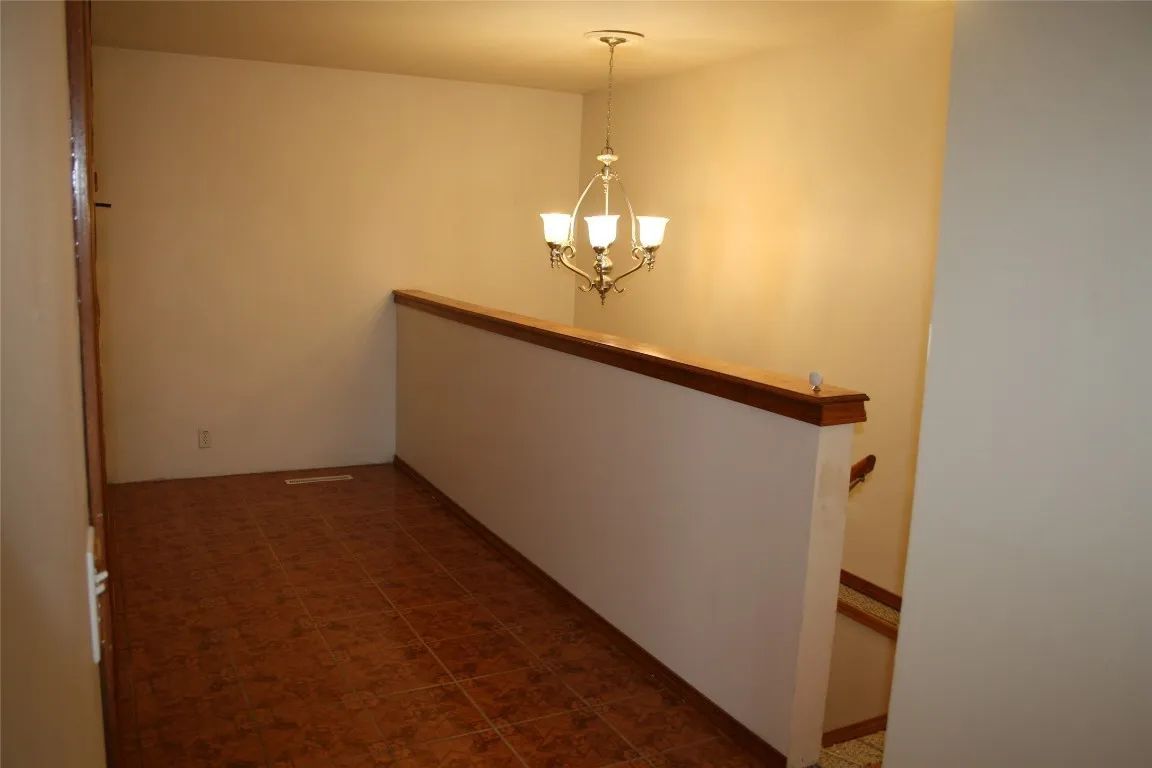 A hallway landing with brown wood-look flooring, a partial wall with a wood railing, and a hanging chandelier.