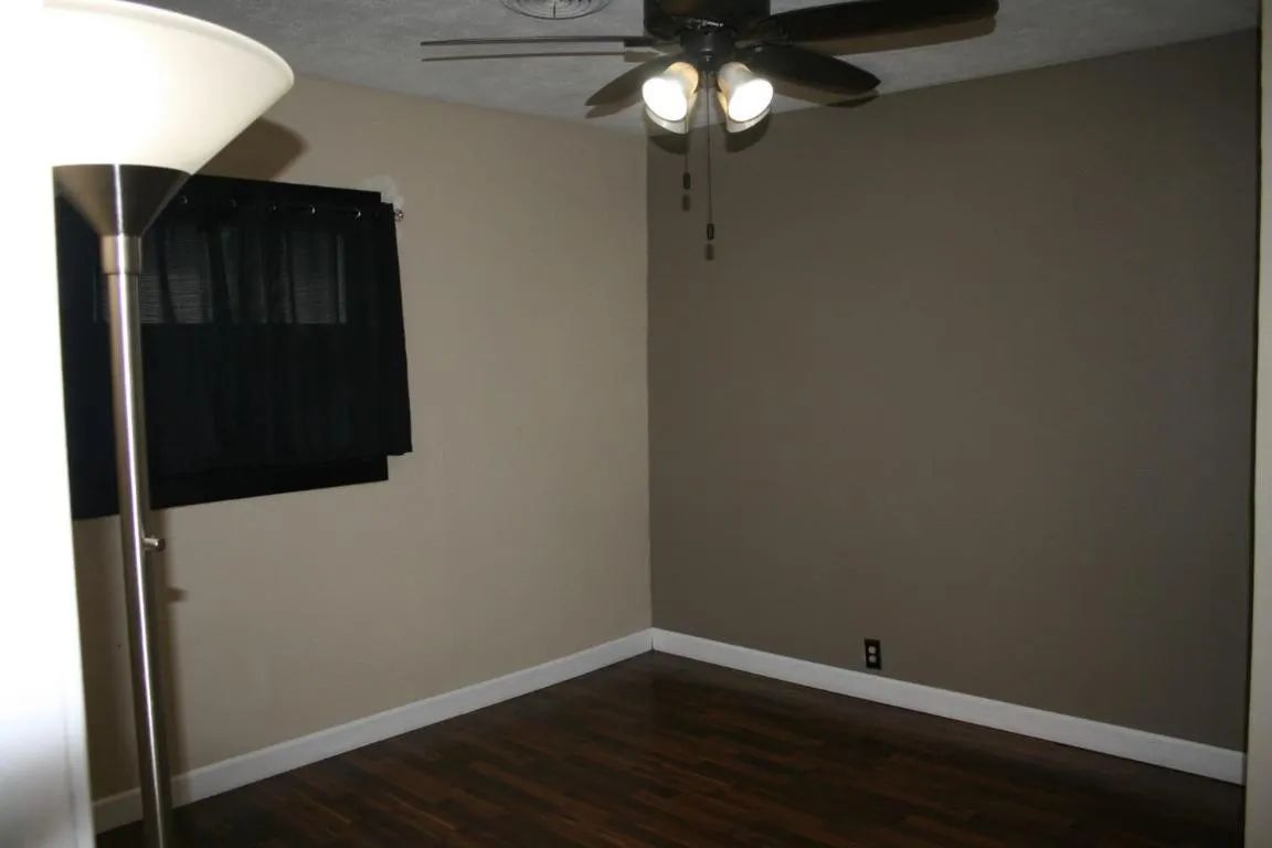 An empty bedroom with dark wood floors, a ceiling fan, and light beige and taupe walls, illuminated by a tall floor lamp.