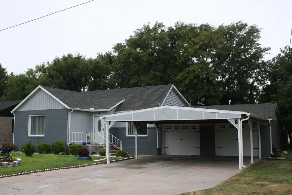 A gray, single-story house with a white carport and matching garage doors, set against a backdrop of mature trees.