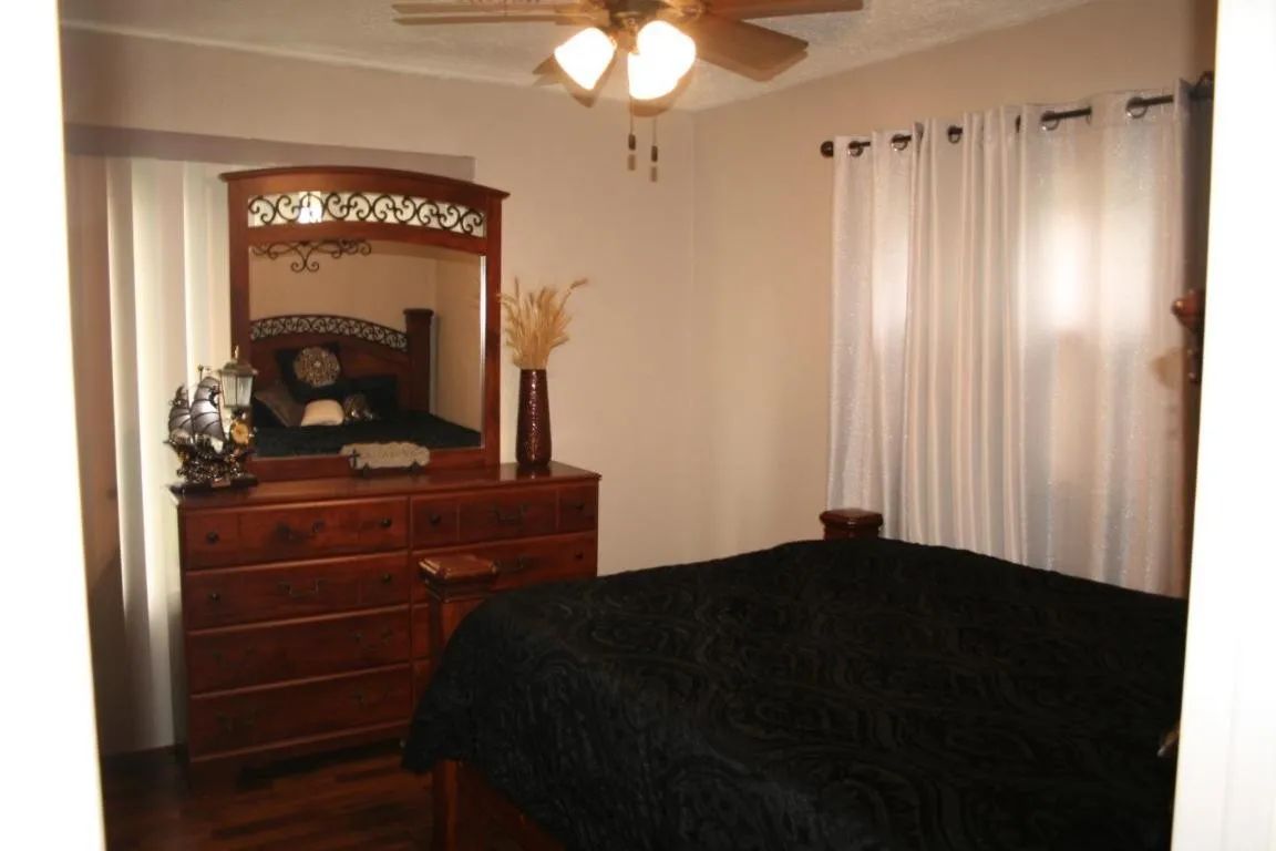 Bedroom with a dark wooden dresser, mirror, a bed with a black comforter, and white curtains covering a window.