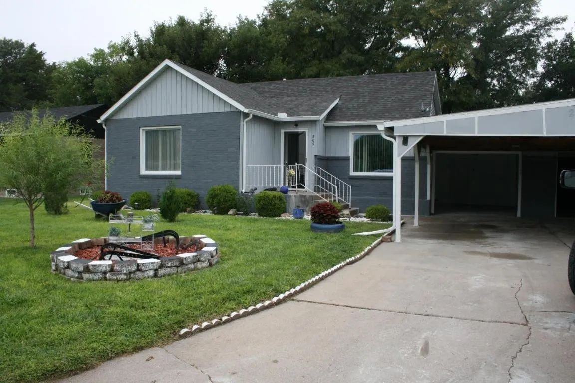 A gray, one-story house with a white-trimmed carport, a stone fire pit in the front yard, and scattered landscaping.