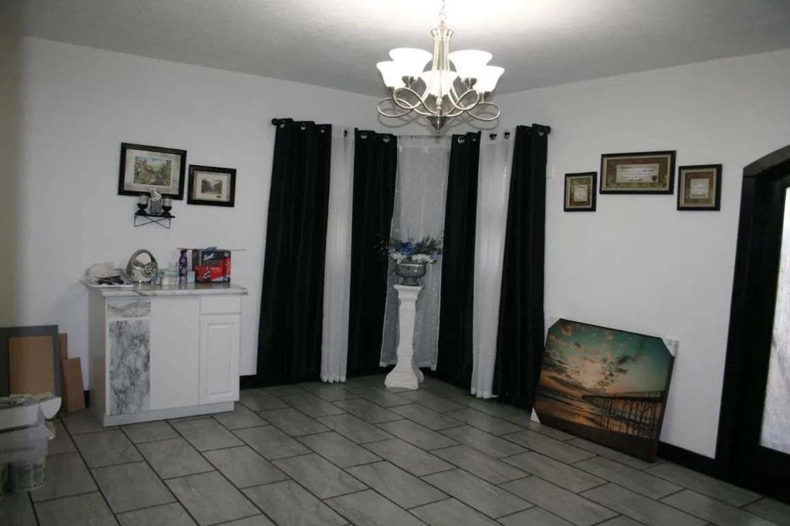 A room with white walls, tiled floors, a chandelier, black and white curtains, framed wall art, and a small cabinet.