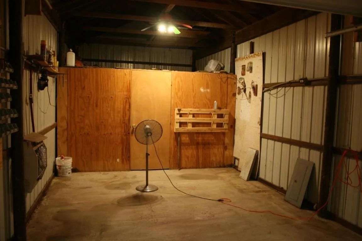 A stand fan sits in the center of a dimly lit garage with wooden walls and a concrete floor.
