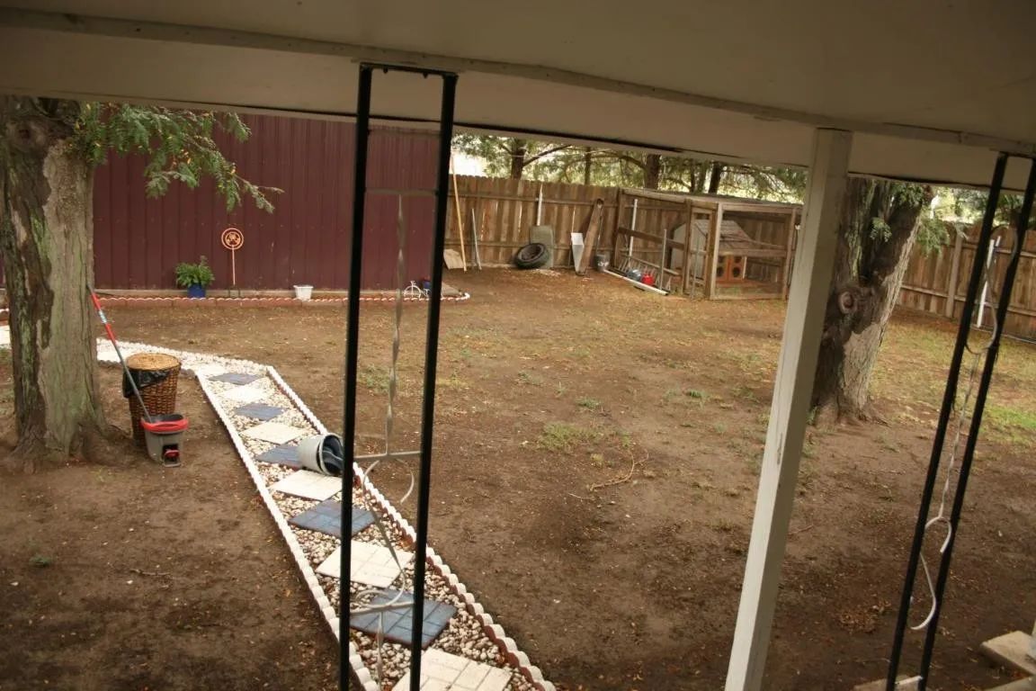 A yard view from a covered patio, showing a dirt ground, a stone pathway, trees, and a wooden coop in the background.