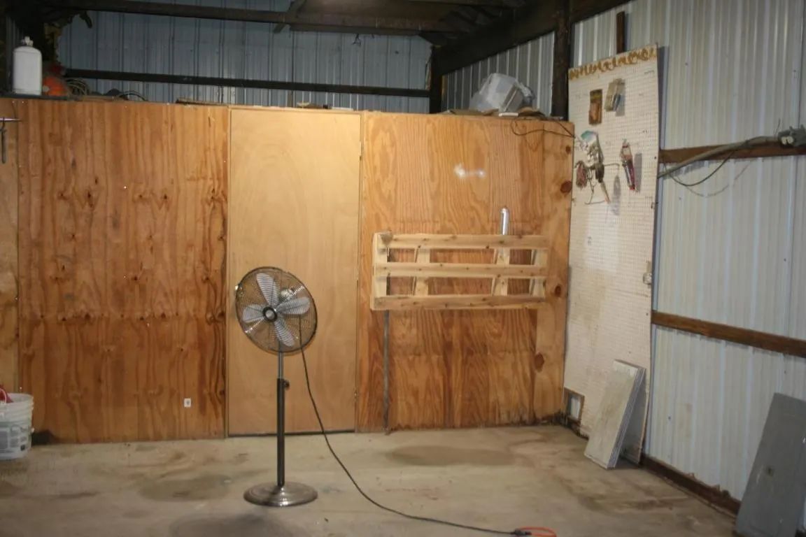 A standing fan in a workshop with plywood walls, a small wooden rack, and exposed metal siding.