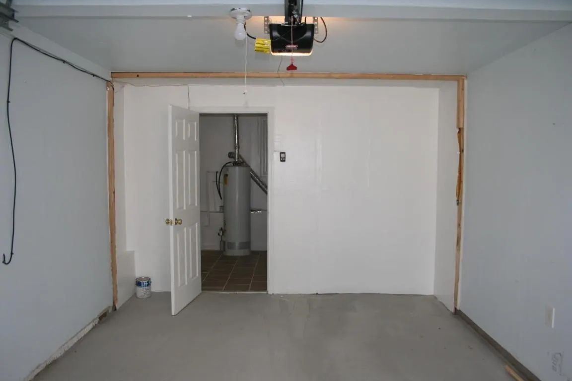 An unfinished garage interior with white walls, concrete flooring, and an open door revealing a water heater.