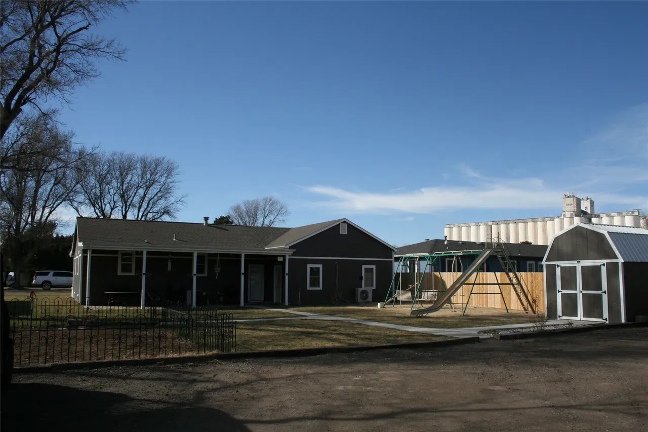 A dark grey house with a porch, a yard with a playset, and a shed, set against a blue sky with large grain silos behind.