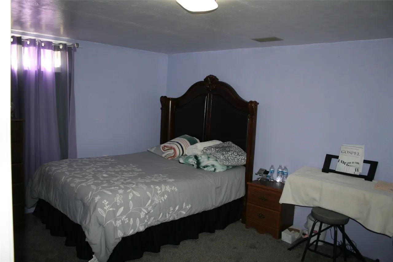 A bedroom with light purple walls features a bed with a dark wooden headboard, a bedside table, and a small music stand.